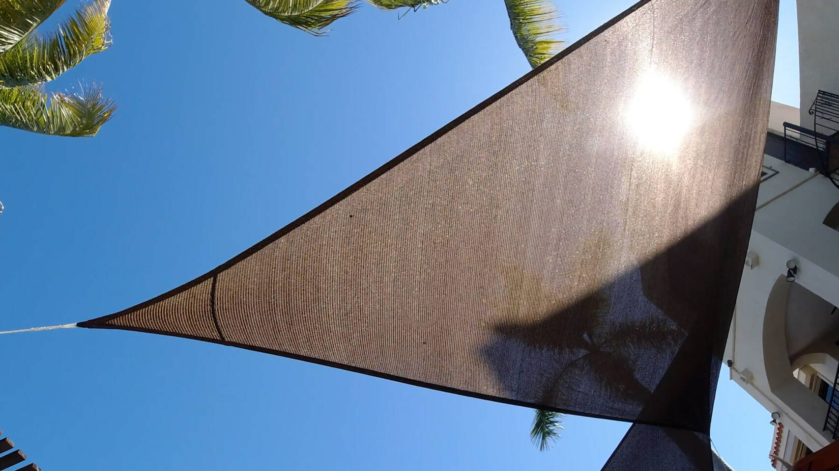 Sunlight filters through a brown shade sail stretched taut against a clear blue sky, with palm fronds and a building visible.