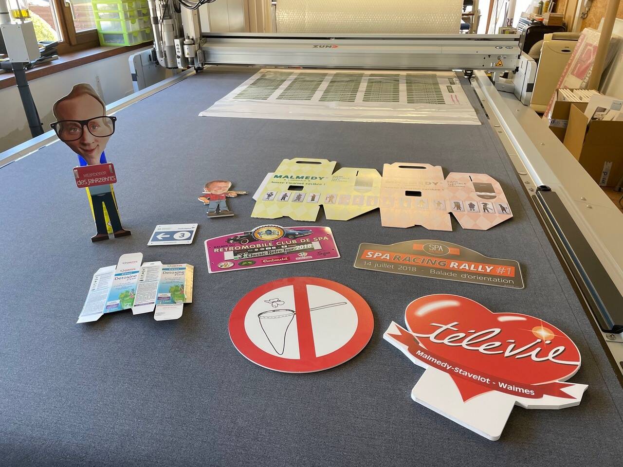 Promotiemateriaal met kartonnen figuren en stickers voor Malmedy, Spa Racing Rally en T&eacute;l&eacute;vie op een grijze tafel.