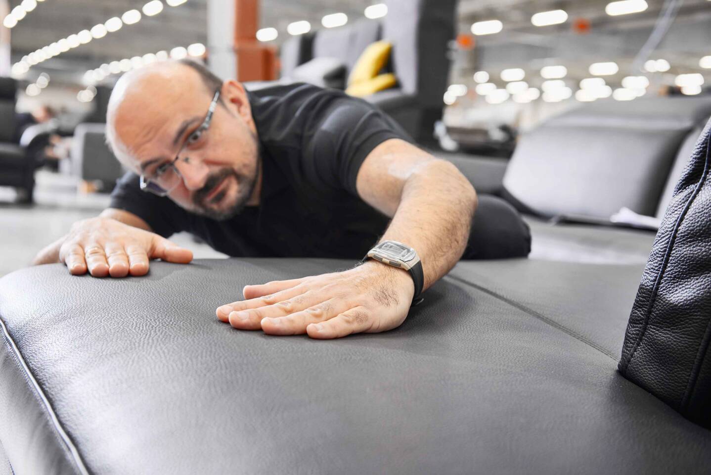 Ein Mann mit Brille und schwarzem Hemd untersucht aufmerksam die Oberfläche eines grauen Ledersitzes. Ein Mann mit Brille und schwarzem Hemd untersucht aufmerksam die Oberfläche eines grauen Ledersitzes.