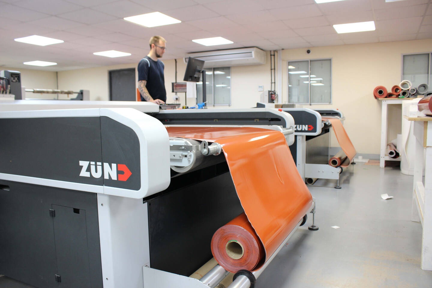 A Z&uuml;nd cutting system processes a roll of orange material, with a technician working in the background.