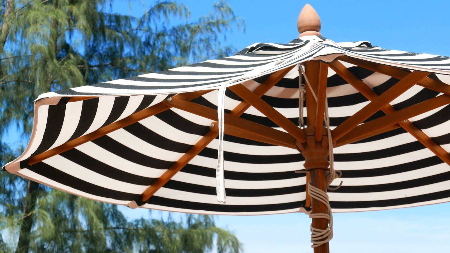 Close-up view of the underside of a black and white striped beach umbrella with wooden ribs against a blue sky and trees.