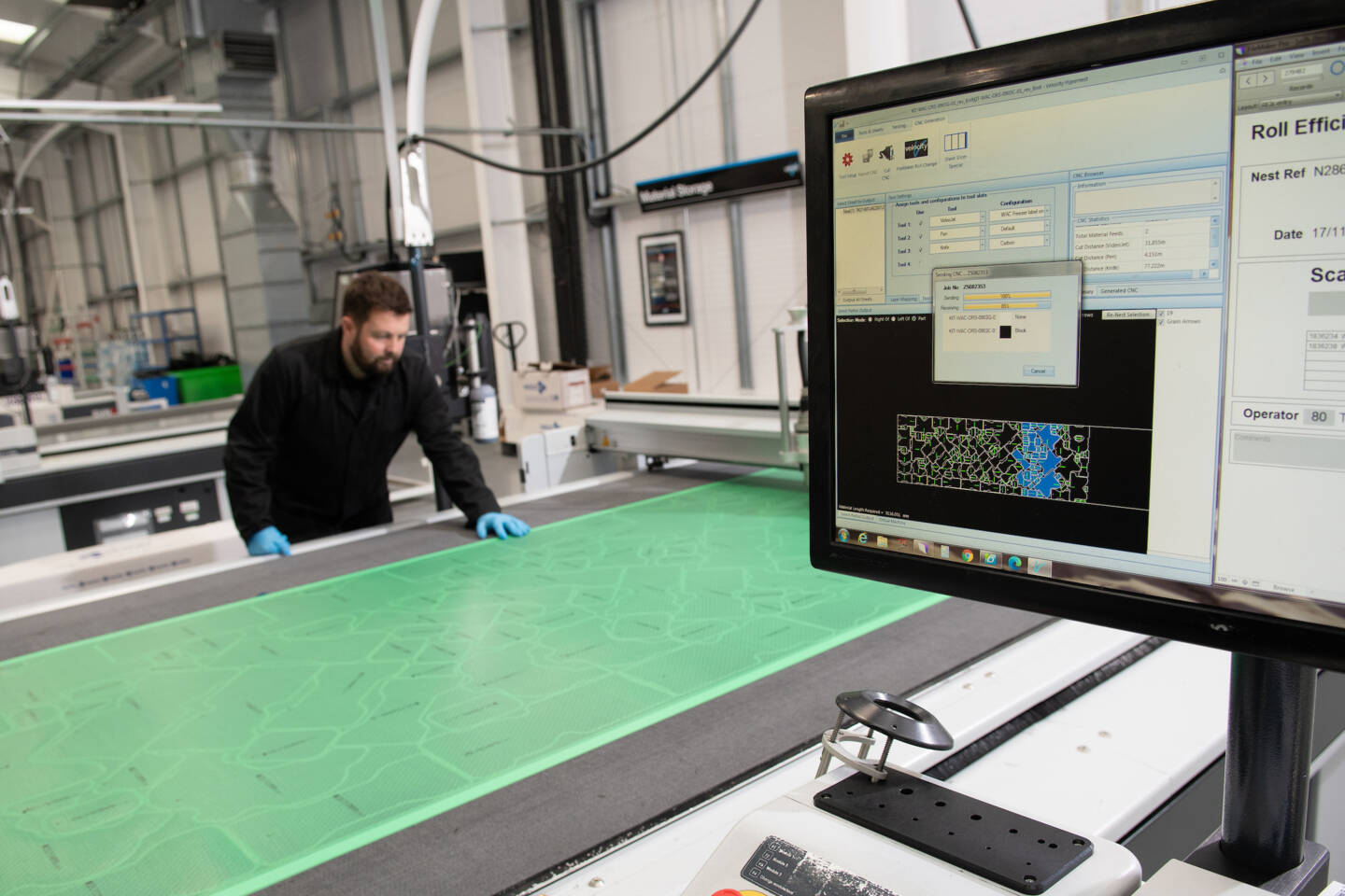Operator in blue gloves inspects a large green material sheet with a pattern on a Zünd cutting system, monitored on a computer screen. Operator in blue gloves inspects a large green material sheet with a pattern on a Zünd cutting system, monitored on a computer screen.