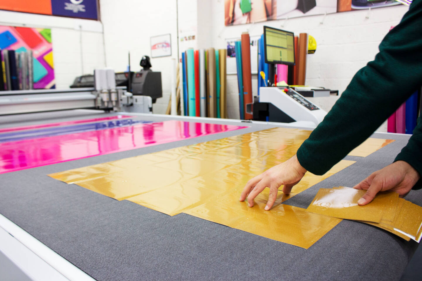 A person's hands place gold material onto a cutting mat next to pink material, with a Zünd cutter in the background. A person's hands place gold material onto a cutting mat next to pink material, with a Zünd cutter in the background.