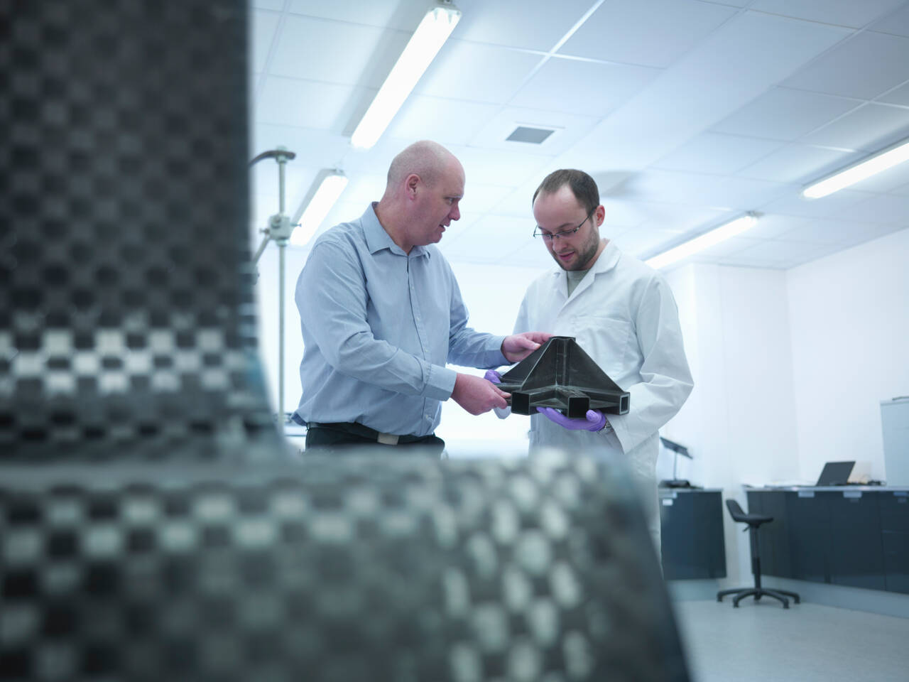 Twee mannen in een laboratorium bekijken een complex zwart composietonderdeel, een in een labjas en de ander in een overhemd.
