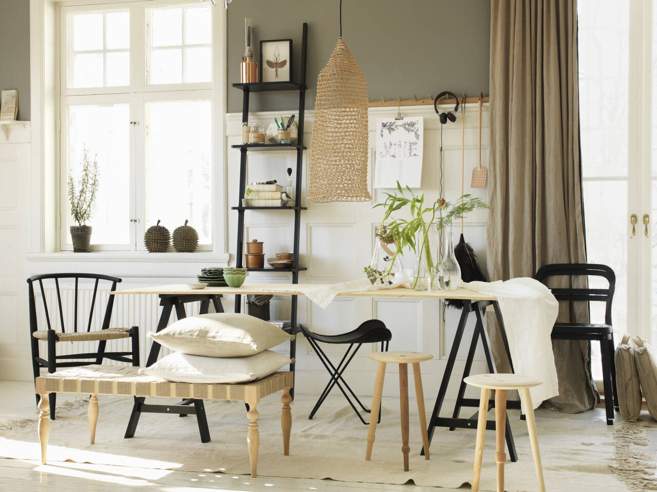 Ein helles, skandinavisches Wohnzimmer mit Holztisch, schwarzen St&uuml;hlen, Kissen, Pflanzen und Regalen, dekoriert mit Heimtextilien.
