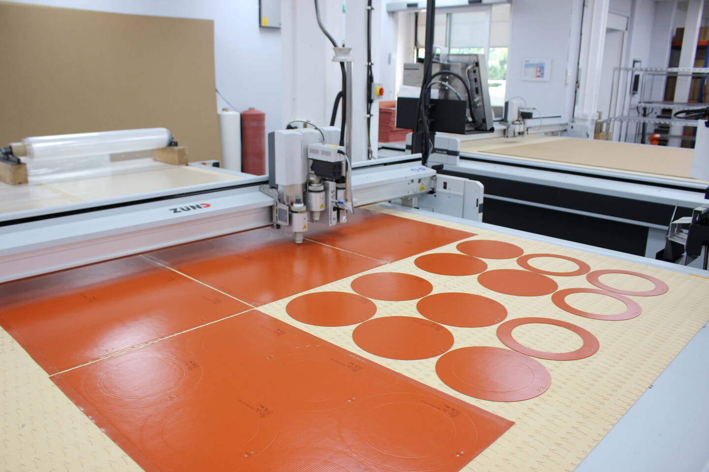 Z&uuml;nd cutting machine precisely cuts circular and oval shapes from orange material on a light-colored surface in a workshop.