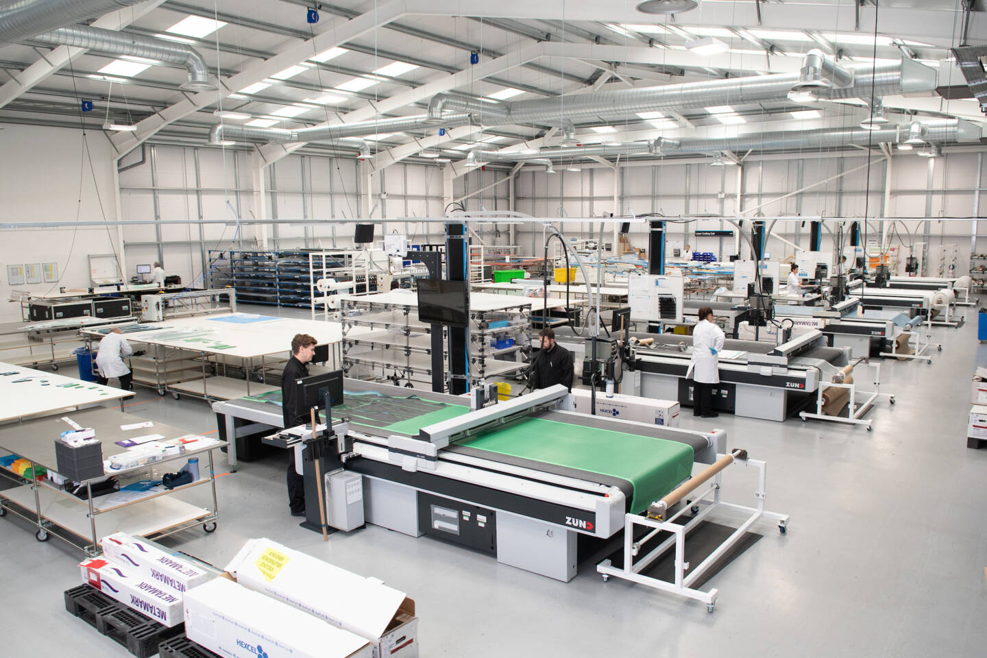 Zünd G3 cutting systems in a modern factory setting, with workers operating the machines and preparing materials for precision cutting. Zünd G3 cutting systems in a modern factory setting, with workers operating the machines and preparing materials for precision cutting.