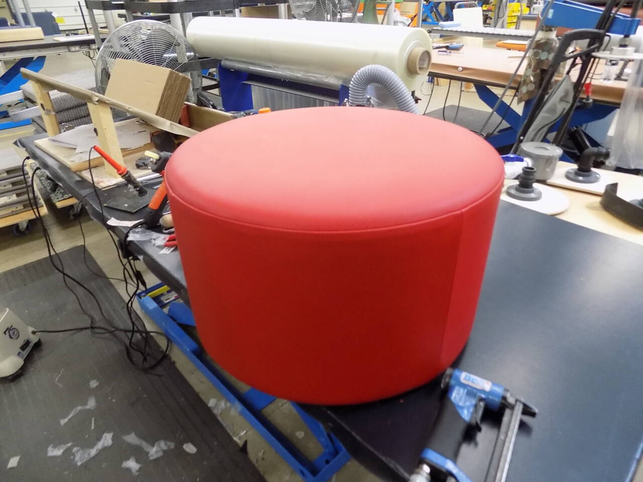 A bright red, cylindrical ottoman sits on a workbench with tools and industrial equipment in the background.