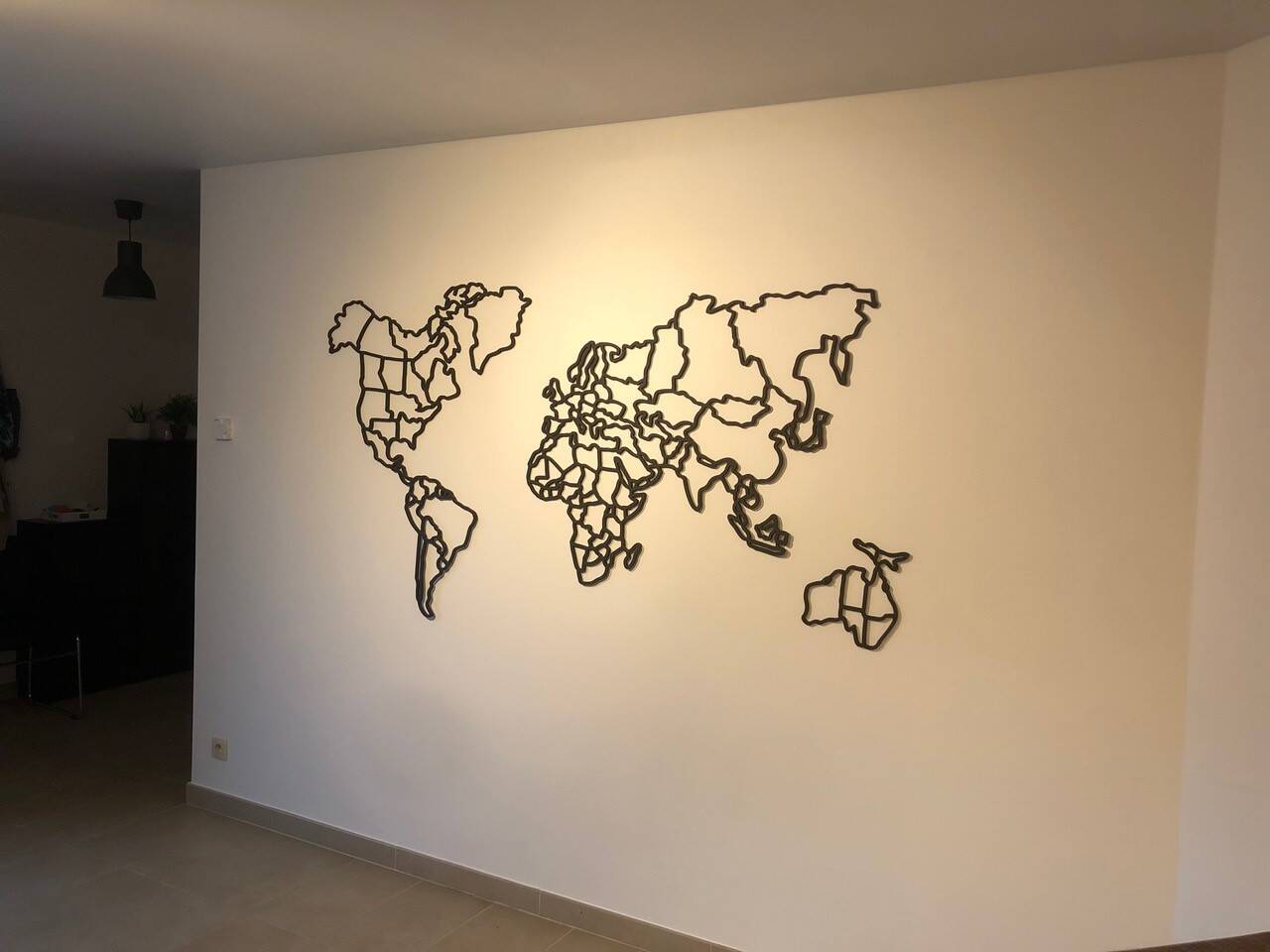 Een zwarte, uitgesneden wereldkaart als decoratie aan een lichte muur, met een lamp en meubels links. Een zwarte, uitgesneden wereldkaart als decoratie aan een lichte muur, met een lamp en meubels links.
