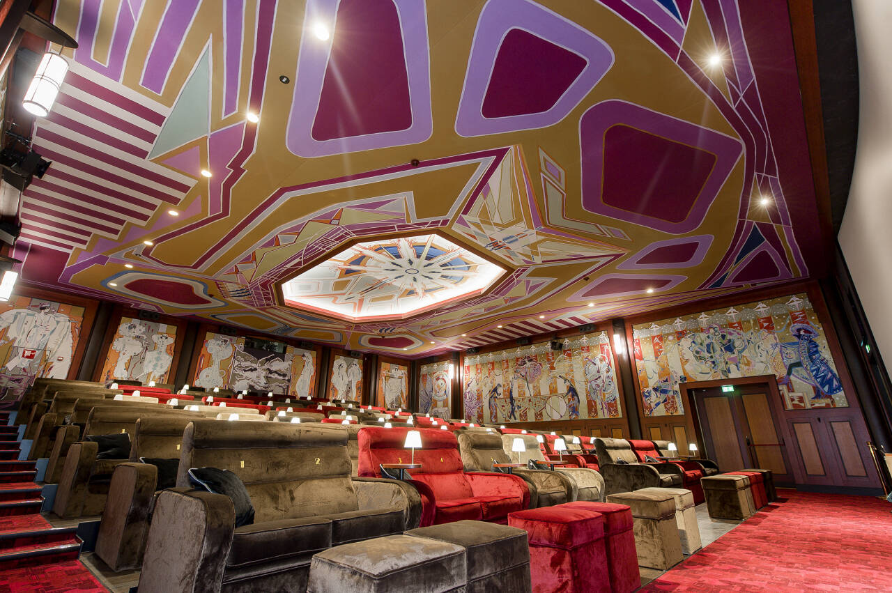 Luxe bioscoopzaal met art deco-plafond, gedetailleerde wandtapijten en comfortabele stoelen met tafeltjes. Luxe bioscoopzaal met art deco-plafond, gedetailleerde wandtapijten en comfortabele stoelen met tafeltjes.