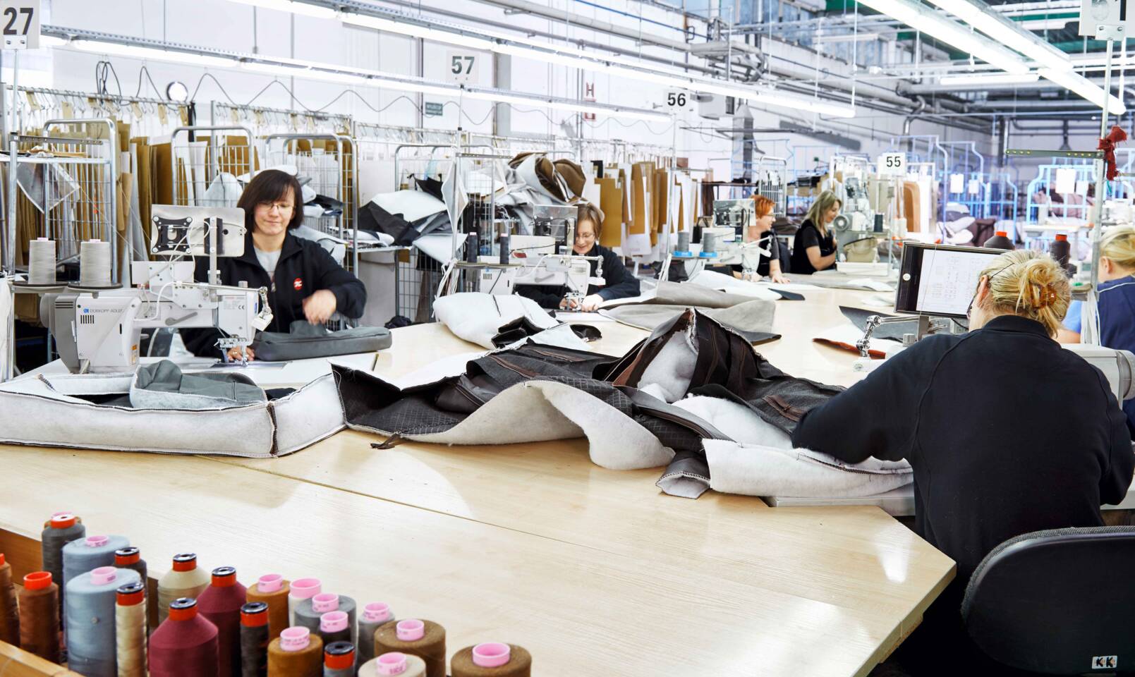 Frauen arbeiten an N&auml;hmaschinen in einer Fabrik, die Leder verarbeitet. Garnrollen sind im Vordergrund sichtbar.