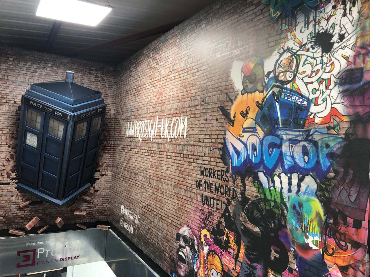 A blue police public call box, resembling the TARDIS, breaks through a brick wall with graffiti art featuring 'DOCTOR' and 'WORKERS OF THE WORLD UNITE'.