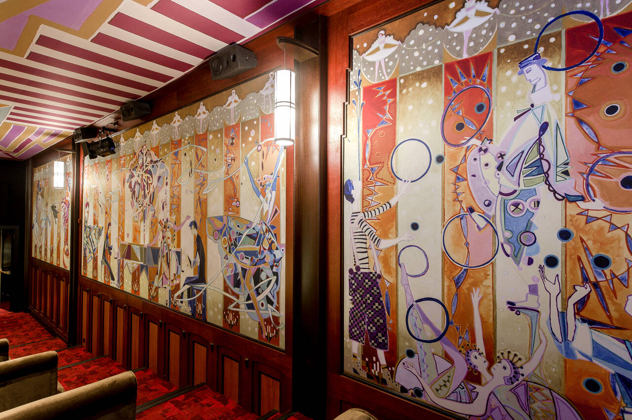 Een muurschildering in Art Nouveau-stijl met figuren en cirkels in een gang met houten lambrisering en rode vloerbedekking.
