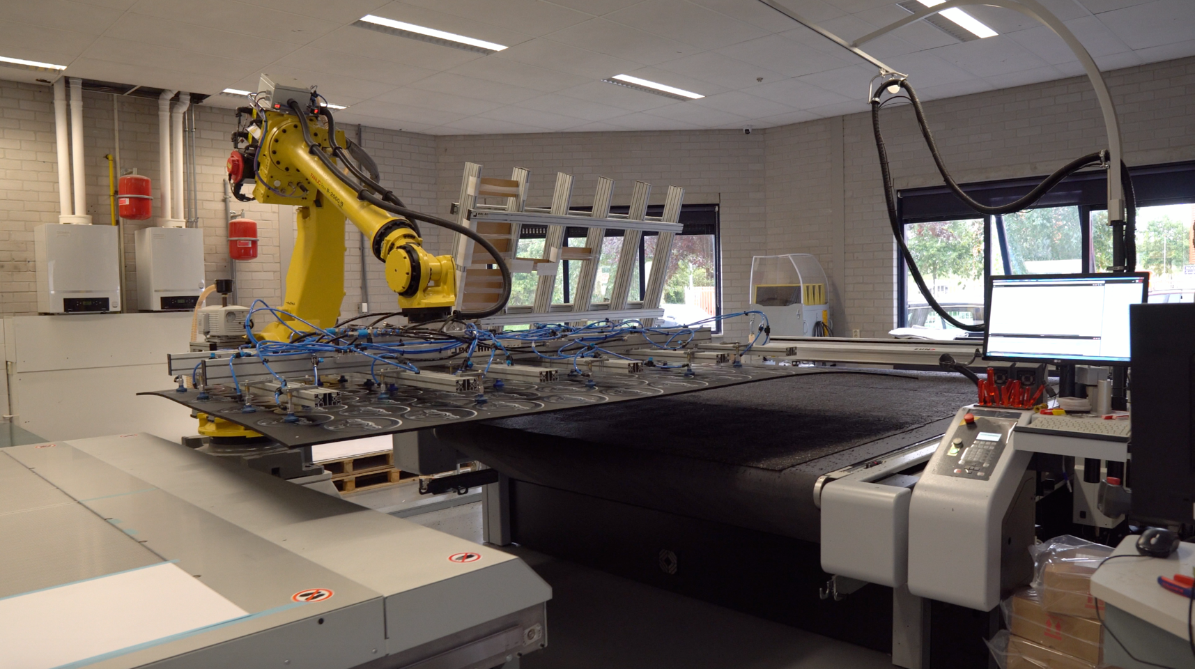 Gele robotarm van Z&uuml;nd Systemtechnik positioneert materiaal op een snijtafel, met computers en ketels op de achtergrond.